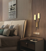 A modern bedroom scene featuring a beige upholstered bed with decorative pillows, a dark wood nightstand, a double-arm wall sconce with illuminated rectangular bulbs, patterned wallpaper, and framed wall art.