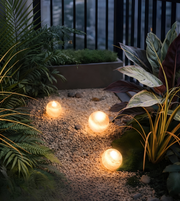 Three glowing spherical garden lights illuminating a landscaped garden path surrounded by lush greenery at dusk.