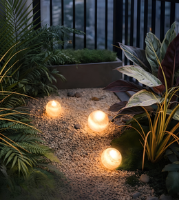 Three glowing spherical garden lights illuminating a landscaped garden path surrounded by lush greenery at dusk.