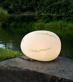 A glowing oval outdoor lamp with a textured, stone-like surface sits on a dark concrete ledge next to a body of water, with green trees in the background.
