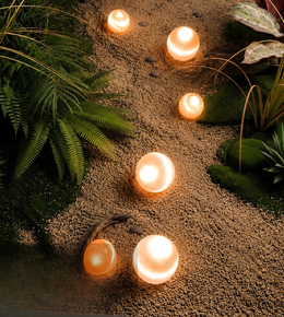 Five glowing spherical solar path lights arranged along a gravel garden path surrounded by green ferns and plants.