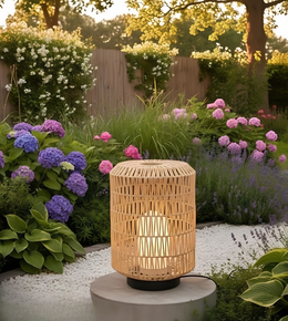 A cylindrical woven bamboo or wicker garden light with a warm glowing bulb inside, placed on a round pedestal in a beautifully landscaped garden with purple and pink hydrangeas, white gravel, and a wooden fence in the background.