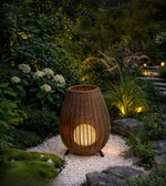 A woven wicker outdoor lantern with a warm glowing light inside, placed on a white gravel path in a lush garden setting at dusk.