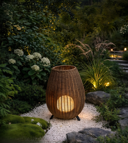 A woven wicker outdoor lantern with a warm glowing light inside, placed on a white gravel path in a lush garden setting at dusk.