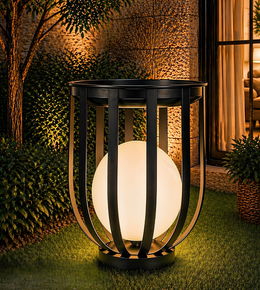 A modern outdoor garden light with a glowing white spherical globe inside a black metal cage, placed on green grass in a landscaped garden setting at night.