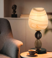 A table lamp with a glowing marbled glass shade and dark sculptural base, placed on a small round table beside a chair in a softly lit interior setting.