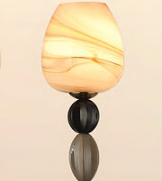 A modern wall sconce featuring a warm, glowing teardrop-shaped glass shade with cream and amber swirls, mounted on a dark metallic stem with a reflective base against a light beige background.