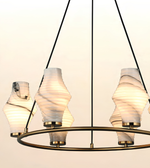A modern multi-light chandelier with four sculptural, ribbed glass shades in a wavy, spiral design. The shades are illuminated with a warm glow, suspended from a circular brass ring with black hanging cords against a plain, light background.