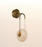 A modern wall-mounted light fixture with a round white and beige marbled glass shade, suspended by a curved brass-toned metal arm from a circular wall plate, shown against a soft warm gradient background.