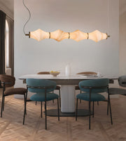 A modern dining room scene featuring a long horizontal pendant light with a wavy, segmented beige shade hanging above a white oval dining table with teal upholstered chairs.