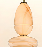 A close-up of a sculptural pendant light with a warm, glowing marbled glass shade in cream and amber tones, featuring a brass-toned metal top and bottom.