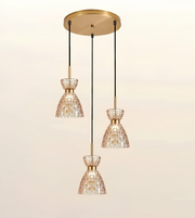 A three-light linear pendant ceiling fixture with a brushed gold canopy and downrod, black cords, and three tapered mosaic glass shades in a warm cream and gold tone, shown against a plain light beige background.