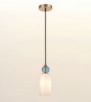 A modern single-light pendant fixture with a round gold ceiling canopy, a black cord, a small blue faceted glass accent, and a frosted white glass shade, shown against a plain off-white background.