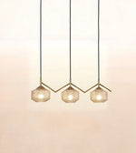 Three modern pendant lights with faceted glass shades and gold-toned geometric metal frames, suspended by black cords against a warm off-white wall.