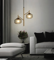 A modern two-light branch pendant lamp with textured gold-finished metal arms and clear glass shades, hanging in a contemporary living room above a white sofa and ottoman.