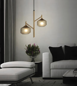 A modern two-light branch pendant lamp with textured gold-finished metal arms and clear glass shades, hanging in a contemporary living room above a white sofa and ottoman.