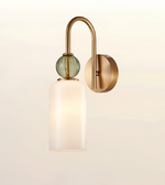 A modern wall-mounted light fixture with a curved brushed brass arm, a round green glass accent, and a frosted white cylindrical glass shade, shown against a plain light background.