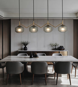 A modern dining room featuring a white marble dining table surrounded by gray upholstered chairs, under four textured glass pendant lights with gold-toned zigzag metal frames.