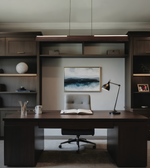 A modern home office setup featuring a dark wood desk and matching built-in shelving, with a gray tufted office chair in front of a framed landscape painting. A black desk lamp sits on the right side of the desk.