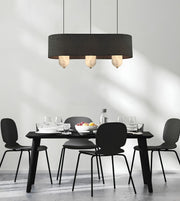 A modern dining room scene with a black rectangular dining table and four black chairs under a large black cylindrical pendant light with three glowing bulbs. The room has a minimalist white wall with soft shadows and a light gray floor.