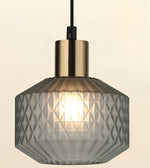 A modern pendant light with a faceted, textured gray glass shade and a brushed brass-finish metal collar, suspended by a black cord against a warm off-white background.