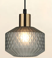 A modern pendant light with a faceted, textured gray glass shade and a brushed brass-finish metal collar, suspended by a black cord against a warm off-white background.