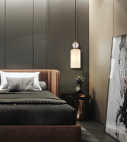 A modern bedroom interior featuring a bed dressed in dark charcoal bedding and white pillows, with a warm brown upholstered headboard and footboard. A black pendant light hangs above a small side table with a vase of flowers, and a large framed abstract print leans against the wall on the right.