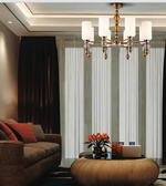 A modern six-light brass chandelier with white cylindrical glass shades hanging in a contemporary living room with a brown sofa, dark drapes, and sheer white curtains.