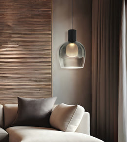 A modern pendant light with a clear glass dome shade and a black cylindrical top, hanging above a beige sofa in a styled living room with wood paneling and curtains.