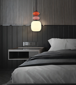 A modern bedroom scene featuring a distinctive pendant light with stacked, rounded glass shades in orange, pink, and white. The light hangs above a bed with gray bedding and a matching dark wood-paneled wall with vertical slats.