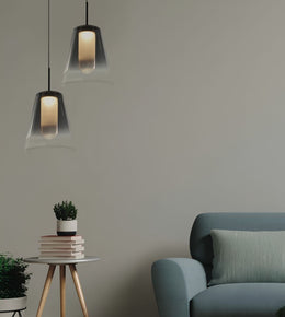Two modern pendant lights with dark metal frames and clear glass shades hang in a minimalist room setting next to a blue-gray armchair and a small side table with books and a plant.