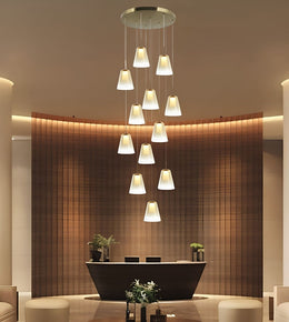 A modern multi-light pendant chandelier with 12 tapered white glass shades hanging in a staggered vertical arrangement from a circular ceiling mount, shown in a luxury lobby setting with a curved wooden slat reception desk.