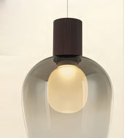 A modern pendant light with a rounded translucent smoky glass shade, a glowing frosted oval bulb inside, and a ribbed cylindrical dark brown cap, shown against a plain off-white background.