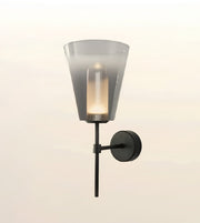 A modern wall-mounted sconce with a translucent smoky gray glass shade, a black metal stem, and a round black wall plate, shown lit against a soft off-white background.