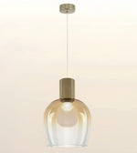 A modern pendant light with a clear ribbed glass shade and a brushed brass cylindrical neck, suspended by a thin cord from a matching ceiling canopy against a warm off-white background.
