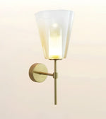 A modern wall-mounted sconce light with a tapered frosted glass shade and a warm brass/gold metal arm and wall plate, shown against a soft off-white background.