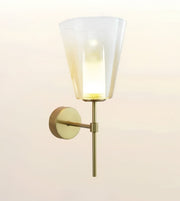 A modern wall-mounted sconce light with a tapered frosted glass shade and a warm brass/gold metal arm and wall plate, shown against a soft off-white background.