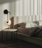A modern wall-mounted black sconce with a circular white shade and curved black arm, installed on a bedroom wall beside a bed and small round side table.