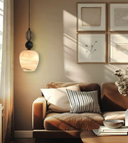 A modern pendant light with a marbled stone-like shade hangs beside a brown leather sofa in a warmly lit living room. The room features a beige wall with framed botanical prints and soft sunlight streaming across the space.