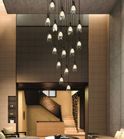 A modern multi-light pendant chandelier with clear glass geometric cone shades and black cords, hanging in a tiered arrangement over a contemporary interior with wood and stone walls.
