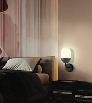 A modern wall-mounted sconce with a glowing white oval shade and a matte dark gray or black base, mounted on a beige wall in a styled bedroom setting next to a bed.