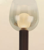 A close-up of a modern floor lamp with a translucent frosted glass bell-shaped shade, a glowing warm white spherical bulb inside, and a dark ribbed cylindrical base against a plain light beige background.