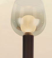 A close-up of a modern floor lamp with a translucent frosted glass bell-shaped shade, a glowing warm white spherical bulb inside, and a dark ribbed cylindrical base against a plain light beige background.