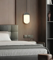 A modern bedroom scene featuring a wall-mounted cylindrical wall lamp with a glowing frosted white shade and a dark gray top, set against a brown vertical slat accent wall above a bed.
