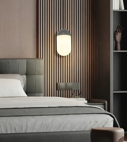 A modern bedroom scene featuring a wall-mounted cylindrical wall lamp with a glowing frosted white shade and a dark gray top, set against a brown vertical slat accent wall above a bed.
