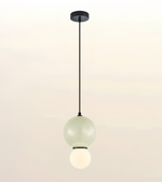 A modern pendant light fixture with a matte black ceiling canopy and cord, featuring two stacked frosted glass globes, one pale green and one glowing warm white, shown against a soft beige gradient background.
