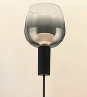 A modern floor lamp featuring a large, translucent smoky gray glass shade and a ribbed black metal stem, shown against a plain light beige background.