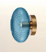 A modern wall-mounted light fixture with a ribbed, translucent blue glass shade and a brushed gold metal wall mount, shown against a soft beige gradient background.