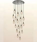 A modern multi-light chandelier with a circular brushed metal ceiling canopy, multiple hanging rods, and twelve leaf-shaped clear glass shades containing warm Edison bulbs, shown against a plain light beige background.