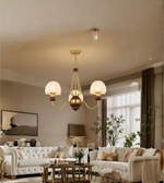 A three-light brass chandelier with white drum shades hangs in a modern living room with cream tufted sofas, a wooden coffee table, and warm ambient lighting.